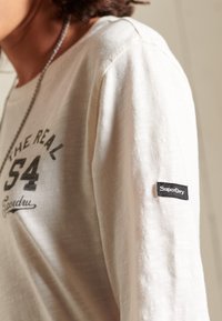 Krämmfärgad långärmad skjorta i mjukt material, med en svart tryckt logotyp och en liten svart SuperDry-etikett på ärmen.