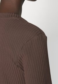 Top en tissu côtelé marron, mettant en avant un gros plan des détails de l'épaule et de la manche, avec des lignes verticales et une texture lisse.