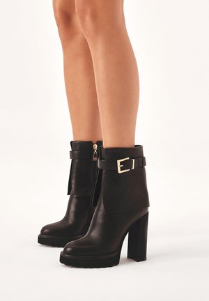 Schwarze Leder-Stiefeletten mit klobigem Absatz, versehen mit einem goldfarbenen Schnallendetail und einem seitlichen Reißverschluss, in leicht konischer Form gestaltet.