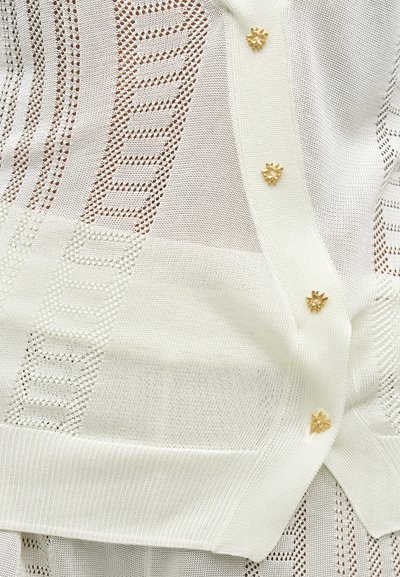 Κρεμ πλεκτή ζακέτα με περίτεχνα ανοιχτά σχέδια και μικρά χρυσά κουμπιά σε σχήμα λουλουδιού μπροστά.