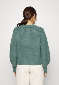 Grön stickad tröja med ribbat mönster, rund halsringning och ballongärmar. Den nedre fållen är åtsittande. Bärs med ljusa byxor.