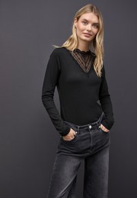 Blonde Frau in schwarzem Spitzen-V-Ausschnitt-Oberteil mit langen Ärmeln und verblassten schwarzen Jeans, die mit den Händen in den Taschen vor einem dunkelgrauen Hintergrund steht.