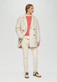 Hellbeige Wollmantel mit doppelten Knöpfen, kombiniert mit einem Korallpullover, cremefarbenen Hosen und Turnschuhen in einem Tan- und Schwarz-Farbschema.