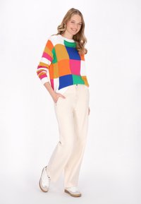 Maglione multicolore con grandi blocchi di arancione, rosa, verde, blu e bianco; abbinato a pantaloni chiari e sneakers bianche.
