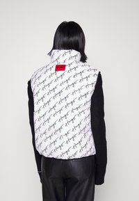Prošívaná vesta v bílé barvě s opakujícím se černým logem "Hugo", vybavená vysokým límcem a červeným detailem loga na zádech, doplněná o černé pletené rukávy.