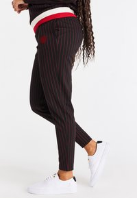 Joggers de rayas negras con franjas rojas, con una banda elástica roja y blanca en la cintura. Combinados con zapatillas deportivas blancas. Tejido ligero con un ajuste entallado.