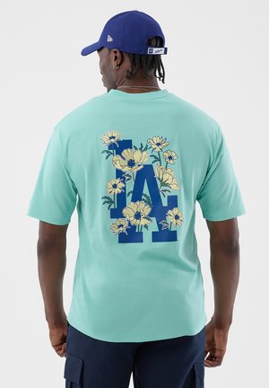 Mężczyzna ubrany w turkusowy t-shirt z dużym niebieskim napisem "LA" i żółtym kwiatowym wzorem na plecach, granatowe spodnie i niebieską czapkę z daszkiem, stojący tyłem.