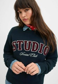 Tumši zila adīta džemperis ar "STUDIO" burgundijas un baltos toņos. Zem džempera valkāts džinsu krekls, ar ribinātām piedurknēm un apakšmalu.