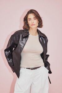 Svart läderjacka med krage, bärs över en ljus beige topp. Matchas med högmidjade vita byxor och ett brunt bälte. Rosa bakgrund.