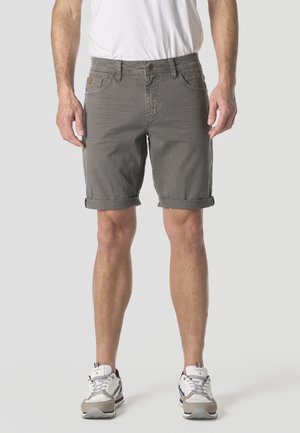 Hombre con pantalones cortos grises hasta la rodilla remangados, zapatillas blancas y camiseta blanca, de pie frente a un fondo claro y liso.