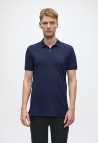 Navy polo shirt with small white pattern, short sleeves, and a collar. Fabric appears smooth and fitted. Worn with dark trousers.