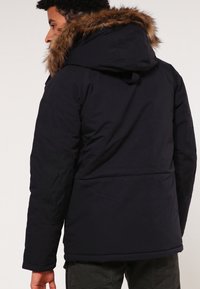 Černá zateplená bunda s kapucí lemovanou kožešinou, texturovanými rukávy a elegantním designem. Tkanina se zdá být odolná a odolná vůči povětrnostním vlivům.