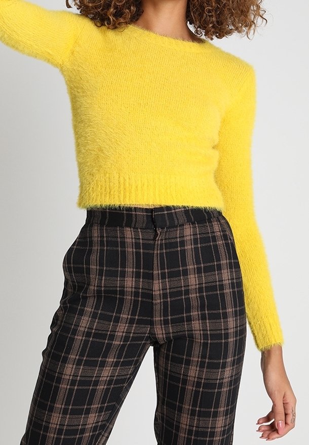Femme portant un pull court duveteux jaune vif à manches longues et un pantalon noir à carreaux taille haute avec des lignes marron et blanches.