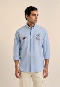 Camisa de botones azul claro con puños enrollados, que presenta un logo de velero bordado y coloridos parches en forma de triángulo en el lado izquierdo del pecho.