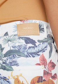 Bílé džíny šortky s živým tropickým vzorem listů a květin. Na zadní straně je kožený štítek v barvě tan s embosovaným textem.