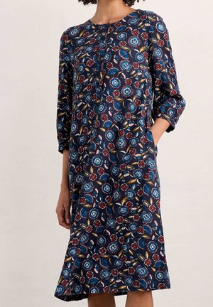 Woman wearing a knee-length navy dress with a multicolored floral pattern and three-quarter sleeves, one hand in pocket.