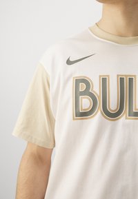 Haut du corps d'une personne portant un t-shirt Nike blanc et beige avec un grand texte « BULLS » à l'avant.