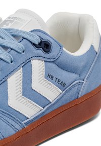 Blå mockasneakers med vita mönster, gummisula och ljusblå snören. Har en "HB TEAM"-etikett och ett unikt snörningssystem.