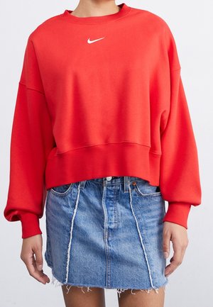 Persoon draagt een rood cropped Nike-sweatshirt en een blauwe denim minirok met rafelige randen en verticale naaddetails.