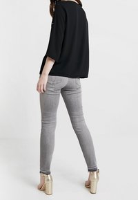 Musta väljä yläosa, jossa on kolme neljänneksen hihaa, yhdistetty kapeisiin harmaisiin farkkuihin ja kultaisiin korokepohjakenkiin, joissa on minimaalinen koristelu ja sileä pinta.