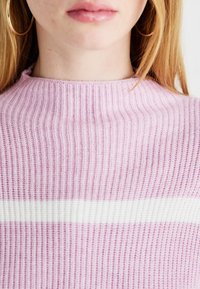 Jasnoróżowy sweter w prążki z białym poziomym paskiem na wysokości klatki piersiowej, z wysokim kołnierzem i miękką fakturą.