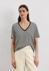 Schwarz-weiß gestreiftes T-Shirt mit V-Ausschnitt, kurzen Ärmeln und einem geknoteten Frontdetail. Kombiniert mit beigen Leinenhosen.