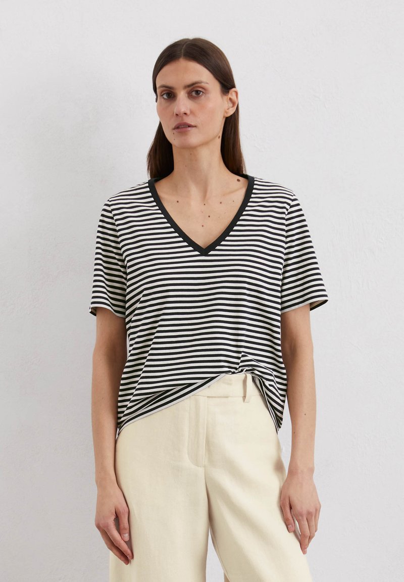 Schwarz-weiß gestreiftes T-Shirt mit V-Ausschnitt, kurzen Ärmeln und einem geknoteten Frontdetail. Kombiniert mit beigen Leinenhosen.