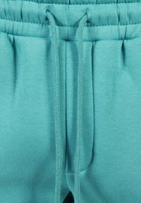 Un pantalon de survêtement bleu-vert avec une ceinture élastique et un cordon de serrage, ainsi que des poches latérales. Réalisé dans un tissu doux au toucher lisse.