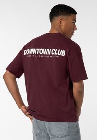 Bordeauxrotes Baumwoll-T-Shirt mit kurzen Ärmeln, einer lockeren Passform und auffälligem weißen Text auf dem Rücken, der "DOWNTOWN CLUB" sowie einen zusätzlichen Satz trägt.