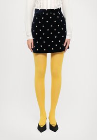 Czarna welurowa mini spódnica z haftem kwiatowym, w połączeniu z żółtymi rajstopami i czarnymi czółenkami na spiczastym obcasie. Biała bluzka z koronkowymi detalami.