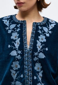 Κοντινό πλάνο ενός ατόμου που φοράει μπλε σκούρο βελούδινο τοπ με γαλαζοπράσινη γεωμετρική κεντητική σε σχήμα λουλουδιού και λεπτομέρειες με χάντρες κατά μήκος της γραμμής του λαιμού και στο μπροστινό μέρος.