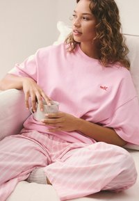 Donna con capelli ricci seduta a gambe incrociate su un divano, indossa una maglietta rosa con la scritta "self." e pantaloni a righe, tiene in mano una tazza di ceramica.