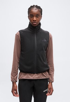 Jeune personne aux cheveux tressés portant un gilet noir zippé par-dessus une chemise marron à manches longues et un pantalon noir sur un fond uni.