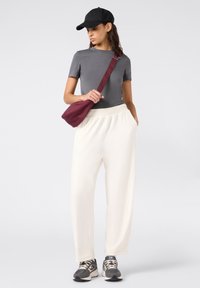 Vrouw in een grijze t-shirt, witte broek, zwarte pet, grijze sneakers en een bordeauxrode schoudertas, staand met één hand in de zak.