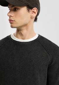 Czarny sweter o teksturowanej powierzchni z białą warstwą pod kolnierzem, wyróżniający się prążkowanym wzorem i rękawami raglanowymi. Model nosi ciemną czapkę.