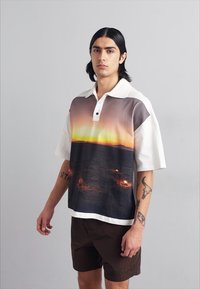 Polo à manches courtes avec un graphisme de coucher de soleil dans des tons chauds, avec un col et des boutons blancs, associé à un short marron.