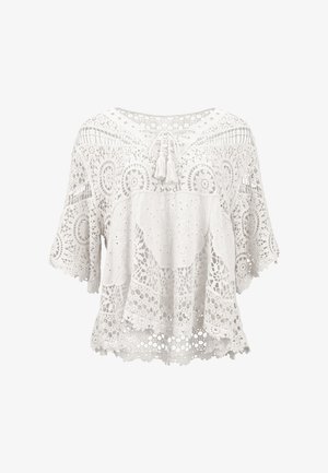 Hvid bluse i blonder med vide ærmer, cirkulære og blomsteragtige mønstre og kvastbånd ved halsudskæringen, vist på en hvid baggrund.