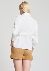 Weißes Langarmhemd mit gerafftem Rücken, ausgestattet mit einem maßgeschneiderten Kragen und weichem Stoff. Kombiniert mit beige Shorts.