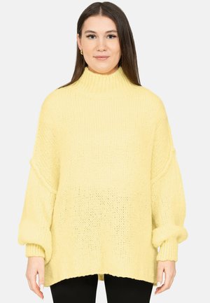 Frau mit langen dunklen Haaren trägt einen lockeren gelben Strickpullover mit hohem Kragen und schwarze Hosen, stehend vor einem schlichten Hintergrund.