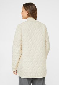 Cream quilted jacket with a diamond pattern, ribbed collar, and long sleeves, featuring a slightly curved hem at the back.