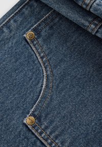 Tejido de mezclilla con un tono azul medio, que presenta una costura curva y un acento de hardware dorado. Los detalles de costura visibles realzan su diseño.