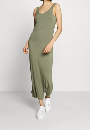 Femme portant une robe maxi sans manches de couleur vert olive associée à des baskets blanches, posant devant un fond clair uni.