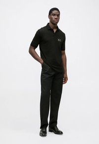 Must värviline polosärk lühikeste varrukatega, millel on väike kuldne logo. Kandke musta pidulikku pükse ja läikivaid musti kingi ning seiske sirgelt.