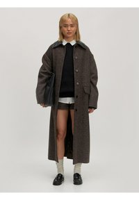 Langer, brauner Mantel aus strukturiertem Stoff mit schwarzem Lederkragen. Ausgestattet mit großen Knöpfen und seitlichen Schlitzen. Kombiniert mit einem dunklen Pullover und Shorts.