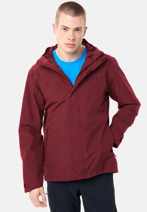 Junger Mann trägt eine kastanienbraune Kapuzenjacke über einem blauen Hemd, steht mit einer Hand in der Tasche vor einem schlichten weißen Hintergrund.