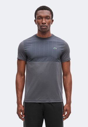 Man draagt een strak grijs sportshirt met korte mouwen en een klein groen krokodillenlogo op de borst, staand tegen een effen achtergrond.