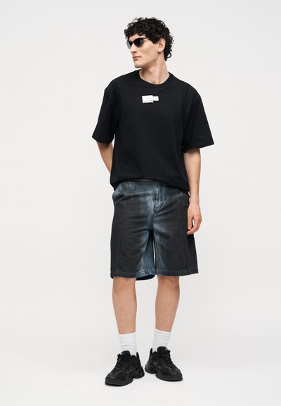 Άνδρας που φοράει μαύρη oversized μπλούζα, σκούρα τζιν σορτς, μαύρα αθλητικά παπούτσια, λευκές κάλτσες και γυαλιά ηλίου, στέκεται με τα χέρια πίσω από την πλάτη.