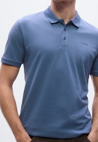 Homme portant un polo bleu uni à manches courtes avec un col et des boutons, avec le logo "HUGO" sur la poitrine.