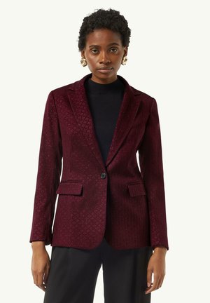 Burgunderfarbener, strukturierter Blazer mit einem Ein-Knopf-Verschluss, Revers und Vordertaschen, kombiniert mit einem schwarzen Oberteil und schwarzen Hosen.
