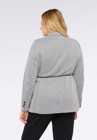Nainen, jolla on vaaleat hiukset, pukeutuneena musta-valkoiseen houndstooth-pukuun ohutmustalla vyöllä, kuvattu takaa, seisten yksinkertaista taustaa vasten.